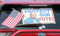 A poster of John Lewis that says "Protect Our Vote" on the back of a car windshield along with an American flag