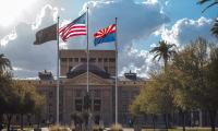 The Arizona state flag with the U.S. flag flying in front of a building with palm trees in the background