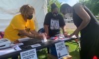 Three women lean over a table at an outdoor booth filling out paperwork