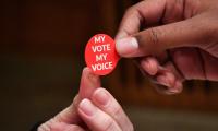 Close up of a person handing a red sticker which says "My Vote, My Voice" to another person.