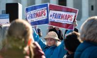 A woman standing in a crowd holds two signs that say "#FAIRMAPS"