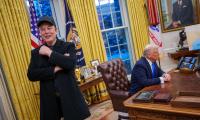 Elon Musk standing up with a smile on his face while Donald Trump sits behind him at his desk in the Oval Office