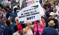 A woman in a crowd holds a sign that reads "And on the 5th day he fired all 17 inspectors general. Why?"