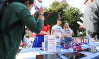 Two smiling people stand behind a table with a sign that says "Register as a voter". Two other people are coming up to the table.