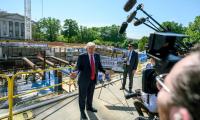 Trump standing in front of a construction site talking to a group of reporters with microphones and cameras.