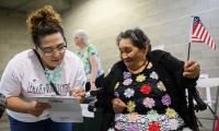A woman lean fills out a paper on a clipboard next to another woman looking on who is holding a small American flag