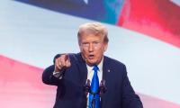 Donald Trump speaking while pointing at the camera in front of a giant American flag
