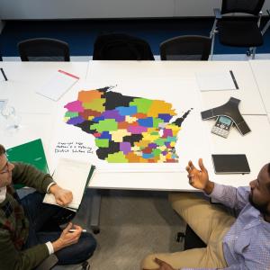A bird's eye view of a map of Wisconsin on a table with district lines drawn and two people sitting next to it discussing.
