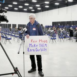A man standing in front of a TV camera holding a sign that says "Fair Maps. Make them earn our vote."