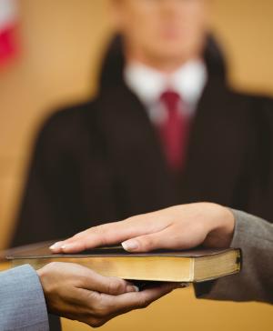 A woman's hand holds a book while another woman places her hand on top. A man in a suit is in the background with an American flag.