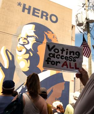 A woman holds a sign which says "Voting Access for All" while standing in front of a large mural of John Lewis.