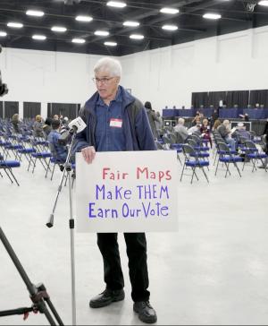 A man standing in front of a TV camera holding a sign that says "Fair Maps. Make them earn our vote."
