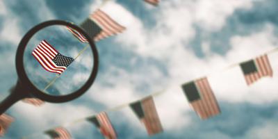 A magnifying glass highlighting one American flag out of a string of them hung up in front of a blue sky with white clouds