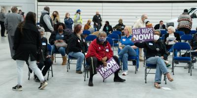 People sit on folding chairs in a gymnasium holding sign that say "Fair Maps NOW!"