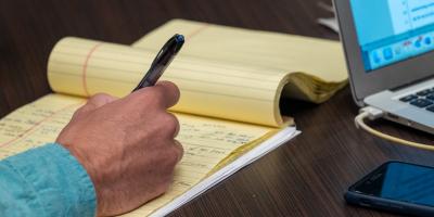 A man's hand writing on a yellow legal pad next to a laptop
