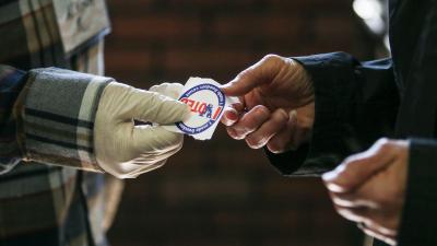 Gloved hand handing an 'I Voted' sticker to another person not wearing gloves