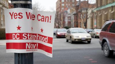 A sign saying "Yes we can! DC Statehood Now!" hanging on a lamppost with traffic going by in the background.