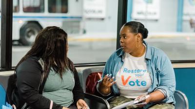 A person wearing a Restore Your Vote t-shirt talking to another person on a bench.