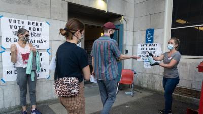 A woman dispenses hand sanitizer to a man and woman outside the open door of a building with "Vote Here" signs on the outside.