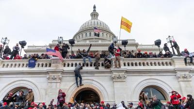 Protestors in a large crowd at the base of the Capitol building as well as climbing up and on top of the wall surrounding it.