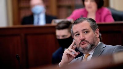 Ted Cruz resting his head on his fingers while sitting at a desk with several people out of focus in the background behind him.