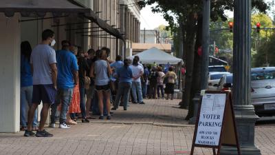 Voters stand in line on a sidewalk. There is a sign about where voters should park. 