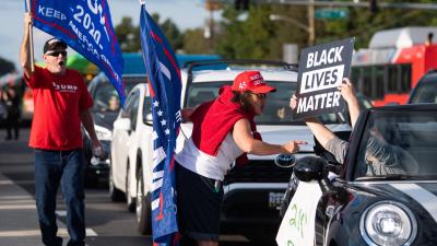 A woman wearing a "Make America Great Again" hat points her finger and yells at a person driving by in a car holding up a "Black Lives Matter" sign.