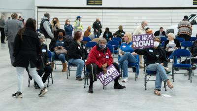People sit on folding chairs in a gymnasium holding sign that say "Fair Maps NOW!"