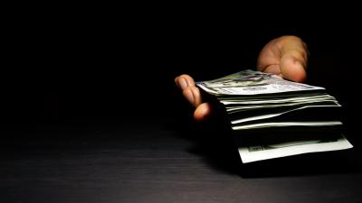 A hand emerges out of the dark holding a stack of $100 bills