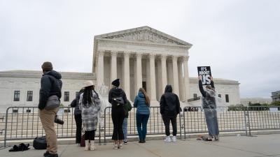 People stand a long a fence in front of the Supreme Court. One woman wearing a poncho holds up a sign which says "It's about US"