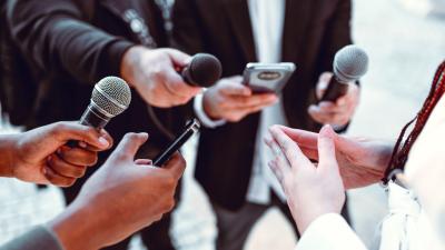 A group of reporters hold up microphones and cellphones, reporters and interviewees faces are obscured.