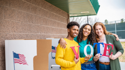 A picture of three people holding letters that spell VOTE. They are standing close together in front of a polling location.