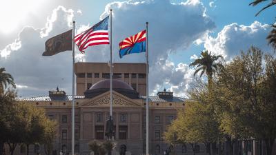 The Arizona state flag with the U.S. flag flying in front of a building with palm trees in the background