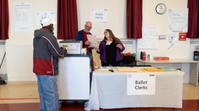 A woman and a man examine a piece of paper while standing next to a machine. A man stands facing them. A sign on the wall says "Ballot Clerks"