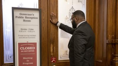 A man stands with his hand against a door. A sign on the doors reads "Governor's Public Reception Room, Closed, Do Not Enter, Meeting in Progress"