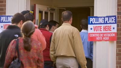 People line up outside a door next to a sign that says "Polling Place, Vote Here"
