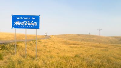 A road sign that says "Welcome to North Dakota" next to a road in the middle of a prairie.