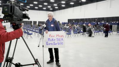 A man standing in front of a TV camera holding a sign that says "Fair Maps. Make them earn our vote."