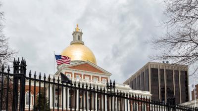 The Massachusetts state house under a cloudy sky with a flag flying and an iron fence in front of it
