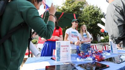 Two smiling people stand behind a table with a sign that says "Register as a voter". Two other people are coming up to the table.