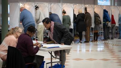 A woman leans over a table to look at paperwork with two other women sitting behind it. In the background is a row a voting booths with voters inside them