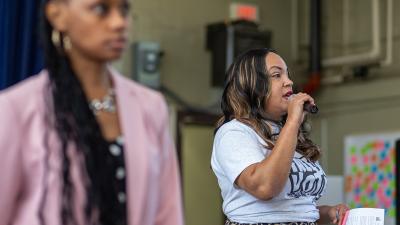 A woman speaking into a microphone with another woman out of focus in the forground.