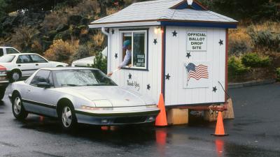 A person in a car hands a ballot to a person leaning out the window of a small hut