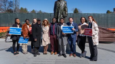 A group of people standing in front of a statue holding signs that say "Maryland needs a voting rights act"