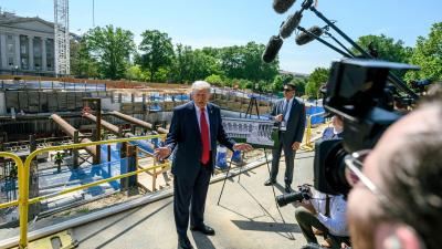 Trump standing in front of a construction site talking to a group of reporters with microphones and cameras.