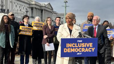 Representative Ayanna Pressley standing at a podium with a sign saying "The Inclusive Democracy Act" in front of the U.S. Capitol with Senator Peter Welch and a group of others standing behind her, some holding signs saying "Voting Rights for All".