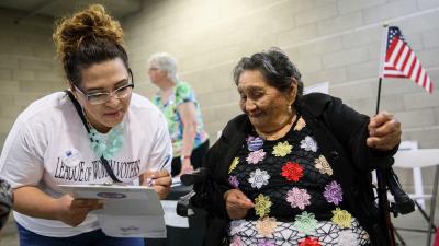 A woman lean fills out a paper on a clipboard next to another woman looking on who is holding a small American flag
