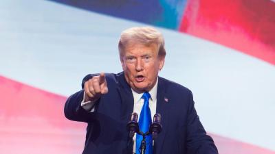 Donald Trump speaking while pointing at the camera in front of a giant American flag