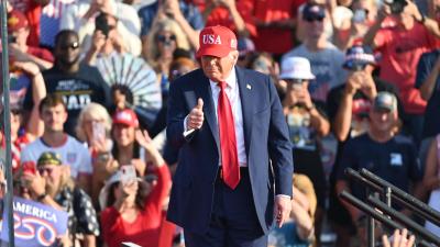 Donald Trump wearing red tie and usa hat giving thumbs up to a crowd of people.