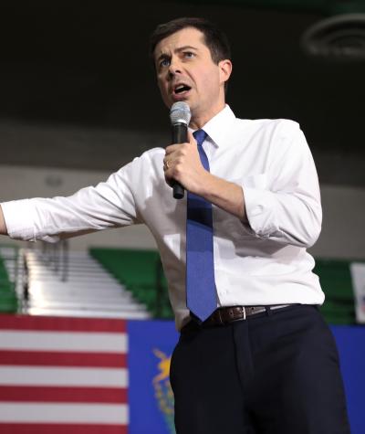 Pete Buttigieg speaking with a microphone on a stage with an American flag in the background